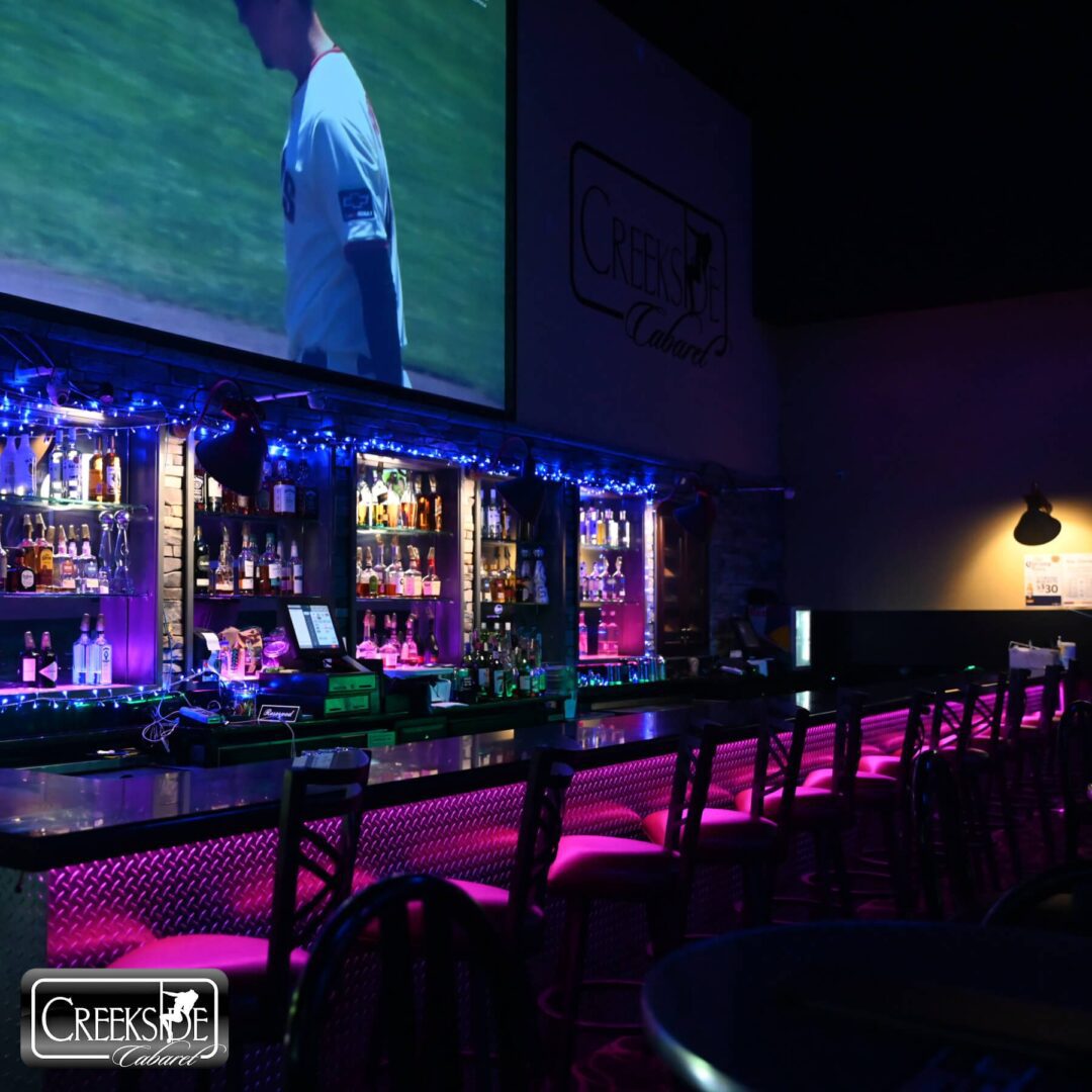 A vibrant bar with purple lighting and a large screen showing a sports game.